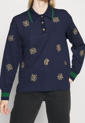 Polo bleu marine à manches longues avec col et poignets rayés verts, orné de blasons brodés en or éparpillés et de boutons sur le devant.