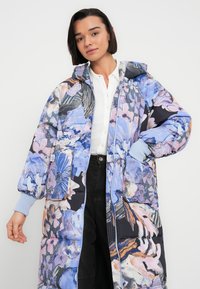 Manteau matelassé à motifs floraux bleu, rose et noir ; doté d'une capuche, de grandes poches et de poignets côtelés ; texture brillante et lisse.