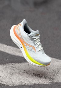 Baskets de sport blanches avec des accents orange et jaune, tige en mesh texturé et semelle en mousse épaisse. Présente un logo rond et des lacets.