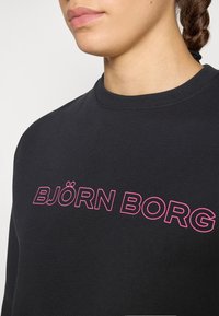 Sweatshirt preto com decote redondo canelado e ousado logotipo em rosa "BJÖRN BORG" atravessando o peito. Tecido suave com textura mínima.