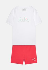 Valge puuvillane T-särk, millel on mündiroheline "diadora" logo ja punane aktsentlipp; koos punaste puuvillaste lühikeste pükstega, millel on valge logo detail.