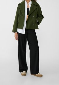 Grön dubbelbröstad peacoat i texturerat tyg, kombinerad med en vit skjorta, svarta vida byxor och beige plattformsskor.