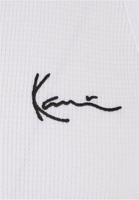 Hvitt ribbemateriale med en svart kursiv "Kani"-logo brodert på overflaten. Teksturen virker myk og noe strukturert.