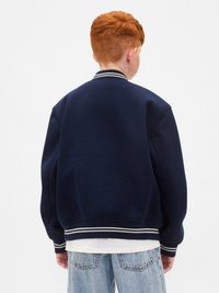 Punapea juuksed laps, kes kannab tumesinist sportlikku jakki ja helesiniseid teksaseid, seisab seljaga välja sirutatuna lihtsa valge tausta ees.