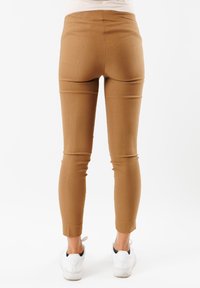 Pantaloni marroni attillati realizzati in un materiale liscio, con vita alta e gambe affusolate. Indossati con sneaker bianche, mostrati da dietro.