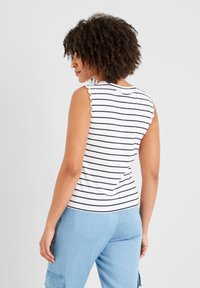 Woman with curly hair wearing a sleeveless white top with black stripes and light blue cargo pants, facing sideways against a plain background.