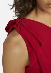 Rode jurk met gestructureerde stof, één schouder met een gestrikte strikdetail, gladde textuur en aansluitende silhoette die de schouder benadrukt.