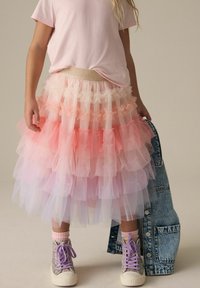 Gonna di tulle stratificata in gradiente rosa e bianco con bordi arruffati, abbinata a una maglietta rosa chiaro e sneakers alte glitterate.