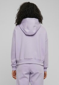 Haut à capuche lilas avec une coupe décontractée, grande capuche arrière, poignets et ourlet côtelés. Fabriqué en tissu lisse au toucher doux. Associé à un pantalon de jogging assorti.