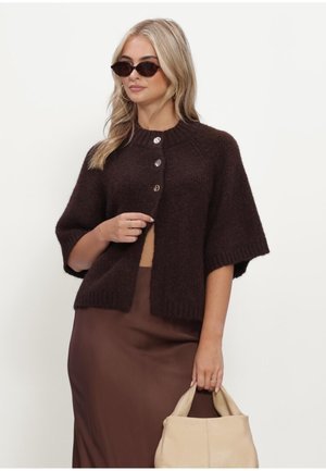 Blonde vrouw met een donkerbruin gebreid vest met knopen, bijpassende bruine rok, zonnebril en houdt een beige handtas vast tegen een witte achtergrond.
