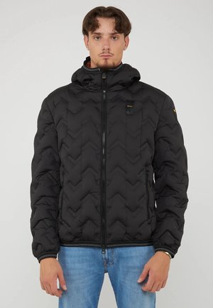 Man draagt een zwart doorgestikt jack met rits en capuchon en blauwe jeans, staand tegen een effen achtergrond.