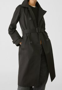 Černý trench coat vyrobený z hladkého materiálu, s límcem, dvouřadým zapínáním na knoflíky, páskem v pase a bočními kapsami.