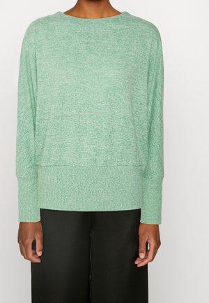 Jasnozielony dzianinowy sweter o luźnym kroju, szerokim dekolcie i ściągaczach przy rękawach, zestawiony z czarnymi spodniami o szerokich nogawkach. Miękka faktura, brak wzorów.