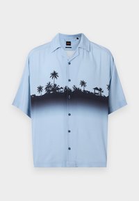 Ανοιχτό μπλε πουκάμισο με κοντά μανίκια και κουμπιά, με μαύρη σιλουέτα από φοίνικες και καλύβες στο στήθος και το πάνω μέρος του κορμού.