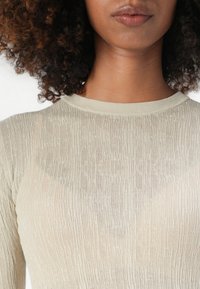 Lys beige langermet topp, ribbestruktur, rund halslinje. Stoffet er gjennomsiktig med vertikale striper som fremhever en kroppsnær silhuett.