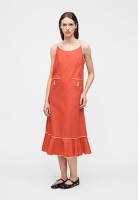 Γυναίκα που φοράει πορτοκαλί μίντι φόρεμα χωρίς μανίκια με κεντήματα λουλουδιών στις τσέπες και στο βολάν στο κάτω μέρος, σε συνδυασμό με μαύρα παπούτσια Mary Jane.