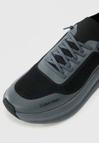 Svart og grå sneaker med et pustende mesh-materiale på overdelen, flate snører, polstret krage og en glatt gummisåle. Merkenavnet er synlig på siden.