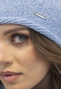 Bonnet tricoté bleu clair avec un motif texturé et un accent logo en argent. Gros plan sur un visage mettant en valeur un maquillage naturel et des traits doux.