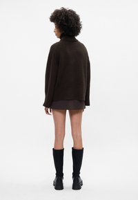 Pārmērīgi liels brūns adīts džemperis ar augstu apkakli, ribotām piedurknēm un malu, apvienots ar krokotu tumšu svārku un melniem zābakiem līdz ceļgaliem.