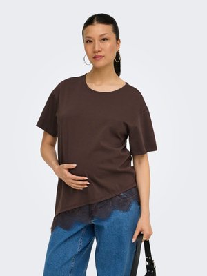 Femeie cu păr închis la culoare prins în coadă de cal, purtând cercei tip cerc, tricou asimetric maro, blugi albaștri și ținând o curea neagră de geantă.