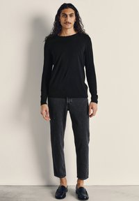 Mladý muž s dlouhými kudrnatými vlasy, oblečený v černém dlouhém tričku, černých džínách a černých mokasínech, stojící před jednoduchou světlou zdí.