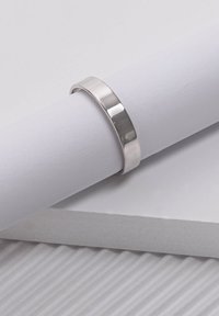 Silver ring with a smooth, polished finish and flat edges, resting on a cylindrical white paper and textured white surface.