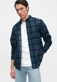 Camisa de cuadro azul marino con una camiseta blanca por debajo, que presenta un cuello abotonado y mangas largas, elaborada con un tejido suave y texturizado.