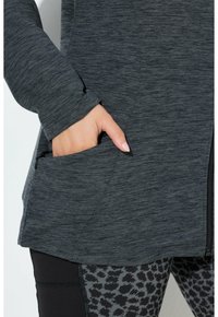 Giacca sportiva grigio scuro con maniche lunghe, tessuto testurizzato e tasche laterali; abbinata a leggings neri con una leggera stampa animale.