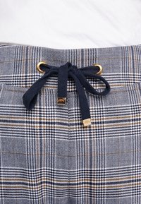Gros plan sur un pantalon à carreaux bleu marine et marron avec un cordon de serrage bleu marine noué en un nœud à la taille à travers des œillets dorés.