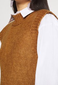 Pull sans manches tricoté marron avec un col côtelé, porté par-dessus une chemise blanche à col. Matière texturée avec une coupe décontractée.