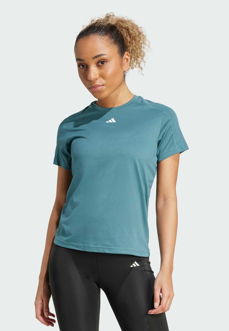 Camiseta deportiva azul verdosa de manga corta, cuello redondo, fabricada con tejido transpirable. Presenta un logo blanco en el pecho, combinada con leggings negros.