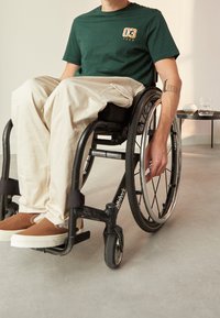 Zelené tričko s krátkým rukávem s potiskem "03 TEAM", béžové volné kalhoty a hnědé slip-on boty; sedící na černém vozíku.