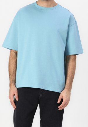 Mand iført ensfarvet lyseblå, oversized kortærmet t-shirt og sorte bukser, stående foran en hvid baggrund.