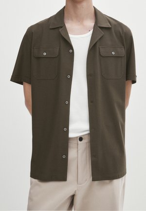 Homme portant une chemise olive à manches courtes ouverte sur un débardeur blanc côtelé et un pantalon beige sur un fond uni.