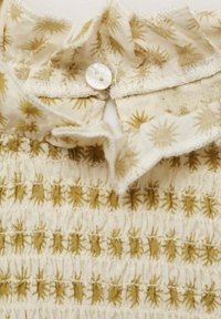 Tessuto di cotone crema con un design smock testurizzato che presenta modelli a raggio dorato. Si chiude con un bottone tondo bianco al collo.