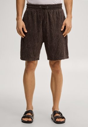 Mężczyzna noszący ciemnobrązowe teksturowane szorty ze ściągaczem w talii i czarne sandały na paski, stojący na jednolitym tle.
