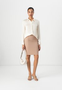 Kremo spalvos marškiniai su mygtukais, prigludusi smėlio spalvos sijonas, sidabriniai šlepetės su dirželiais ir balta apskrito formos rankinė su logotipu. Lygūs paviršiai.