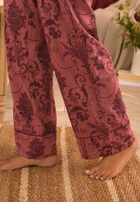 Barfuß steht auf einem gewebten Teppich neben einem Holzmöbel, trägt lockere rote Hosen mit dunklen Blumen- und Weinrebenmustern.