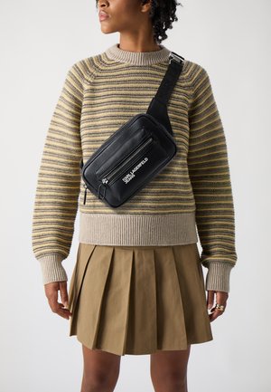 Schwarze Umhängetasche mit mehreren Reißverschlusstaschen und weißem Logo, kombiniert mit einem gestreiften Pullover und einem plissierten khakifarbenen Rock.