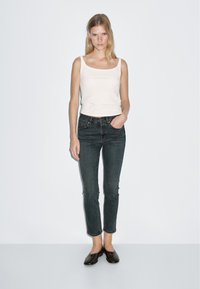 Beige tank top, dark denim jeans with a fitted design, and black flat shoes. Model stands against a plain white background.