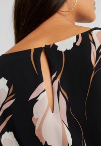 Blouse noire avec un motif floral en rose pâle et blanc. Détail en forme de clé à l'arrière, avec une texture de tissu lisse et une coupe décontractée.