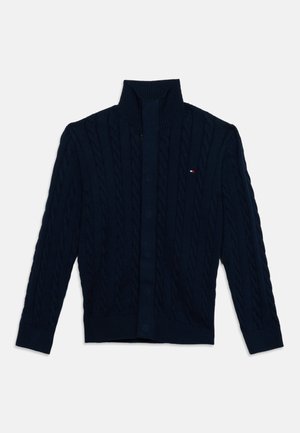 Cardigan en maille torsadée bleu marine avec col montant, manches longues, boutonnage sur le devant et petit logo rouge, blanc et bleu sur la poitrine.