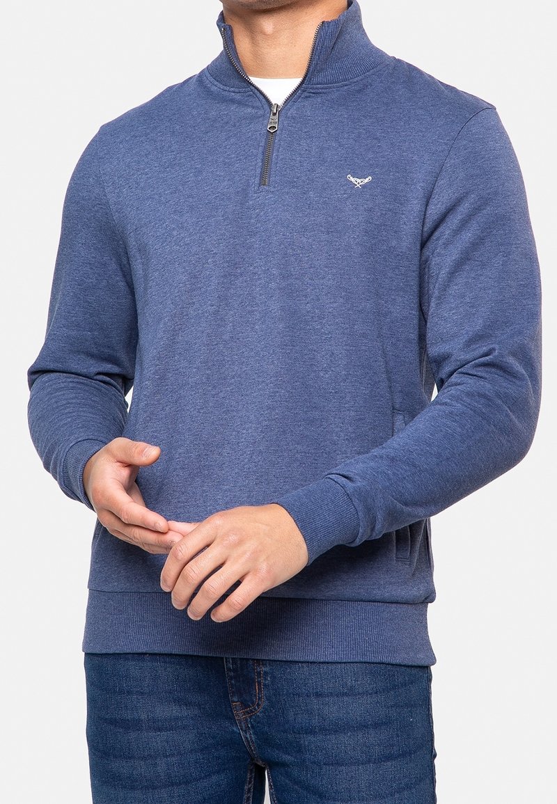 Homme portant un pull bleu à fermeture éclair jusqu'au quart, avec des poignets et un ourlet côtelés, assorti à un jean bleu foncé, mains jointes devant.