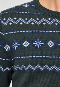 Stickad tröja i mörkgrön bas med vita och blå geometriska mönster. Rund hals, ribbade ärmslut och nederkant. Texturerad yta.