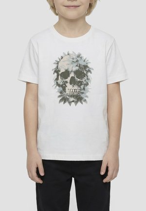 Enfant portant un t-shirt blanc avec un grand motif de crâne entouré de feuilles grises, associé à un pantalon noir, sur un fond neutre.