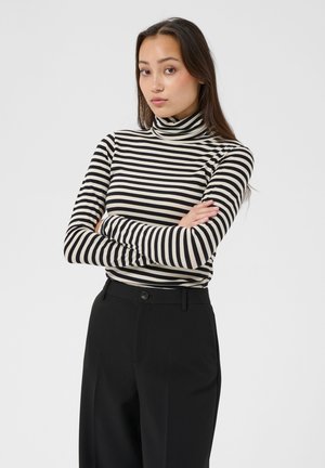 Jeune femme aux longs cheveux foncés portant un col roulé rayé noir et blanc et un pantalon noir, debout les bras croisés devant un fond uni.