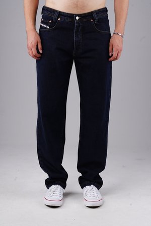 Man wearing dark blue jeans and white sneakers, standing against a plain light gray background.