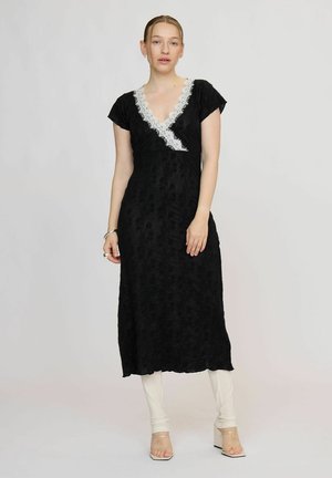 Femme portant une robe en dentelle noire avec une bordure en dentelle blanche au niveau de l'encolure, associée à des leggings blancs et des sandales transparentes à talons hauts.