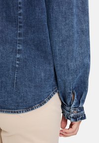 Denimowa koszula w ciemnoniebieskim kolorze z długimi rękawami. Posiada zapinane mankiety i zaokrąglony dół, prezentując gładką fakturę oraz subtelne wyblaknięcia.