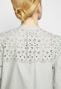 Femme aux cheveux blonds en chignon bas portant un chemisier clair avec une dentelle ajourée complexe au niveau des épaules dans le dos.